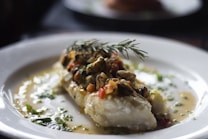 A beautifully plated dish featuring a piece of white fish topped with a savory mixture including mussels, diced red and yellow peppers, and fresh herbs. The dish is garnished with a sprig of rosemary and surrounded by a light, creamy sauce dotted with green herbs, all served on a white plate.