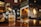A bottle of Johnnie Walker Green Label is prominently placed on a wooden bar counter. A glass with ice cubes is being filled with the same whisky from another bottle. The bar setting is cozy, featuring dim lighting and a brick wall in the background, with several decorative elements such as framed pictures and hanging lights.