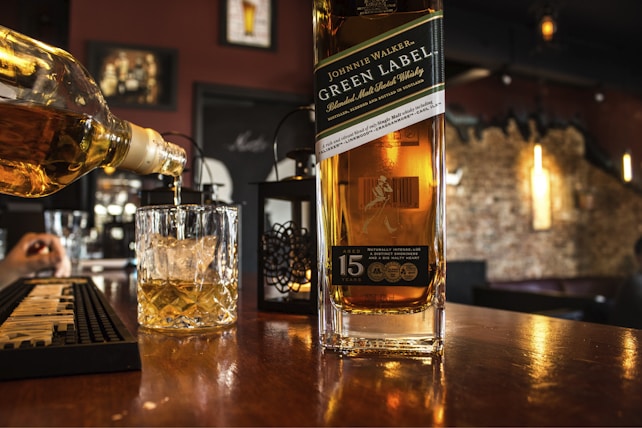 A bottle of Johnnie Walker Green Label is prominently placed on a wooden bar counter. A glass with ice cubes is being filled with the same whisky from another bottle. The bar setting is cozy, featuring dim lighting and a brick wall in the background, with several decorative elements such as framed pictures and hanging lights.