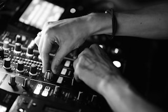 Hands adjusting knobs on a DJ mixing console, with various controls and buttons visible under soft lighting. The focus is on the precision and dexterity required to operate the equipment.