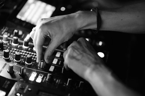 Hands adjusting knobs on a DJ mixing console, with various controls and buttons visible under soft lighting. The focus is on the precision and dexterity required to operate the equipment.