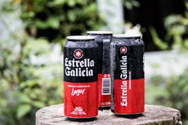 Three cans of Estrella Galicia lager beer with condensation droplets on their surfaces are placed on a wooden stump. The background is blurred with greenery, suggesting an outdoor setting.