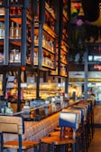 A warm and inviting bar setting with a long, polished counter and lined with wooden bar stools. Shelves above the counter display bottles of various spirits, illuminated by soft, ambient lighting. In the background, the kitchen area is visible, adding a bustling atmosphere to the scene.