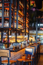 A warm and inviting bar setting with a long, polished counter and lined with wooden bar stools. Shelves above the counter display bottles of various spirits, illuminated by soft, ambient lighting. In the background, the kitchen area is visible, adding a bustling atmosphere to the scene.