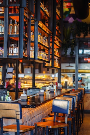 A warm and inviting bar setting with a long, polished counter and lined with wooden bar stools. Shelves above the counter display bottles of various spirits, illuminated by soft, ambient lighting. In the background, the kitchen area is visible, adding a bustling atmosphere to the scene.