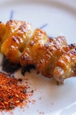 A skewer of grilled chicken with a crispy, golden-brown surface is placed on a plate. Beside it lies a small heap of red and black spices, possibly a mix of chili and sesame seeds.