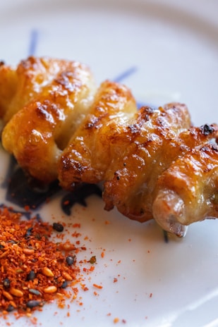 A skewer of grilled chicken with a crispy, golden-brown surface is placed on a plate. Beside it lies a small heap of red and black spices, possibly a mix of chili and sesame seeds.