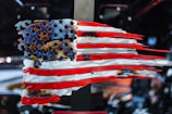 A stylized, abstract representation of the American flag composed of metal with jagged edges. The stars and stripes are artistically distorted, creating a dynamic and rugged appearance. The background is blurred, focusing attention on the flag.