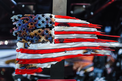 A stylized, abstract representation of the American flag composed of metal with jagged edges. The stars and stripes are artistically distorted, creating a dynamic and rugged appearance. The background is blurred, focusing attention on the flag.