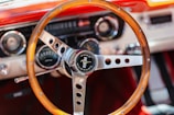 A vintage wooden steering wheel features prominently, with a Ford Mustang emblem at the center. The dashboard, with retro-style dials and controls, is visible in the background, characterized by metallic and wood details. The setting exudes classic car elegance.