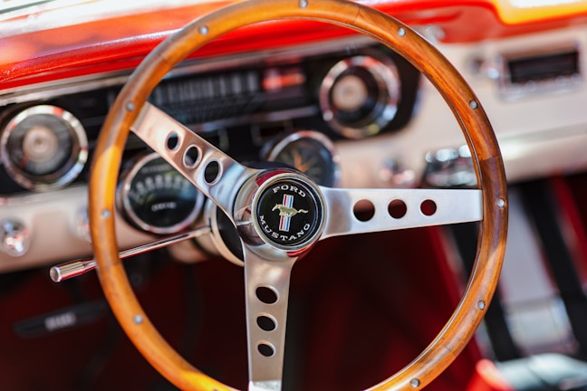 A vintage wooden steering wheel features prominently, with a Ford Mustang emblem at the center. The dashboard, with retro-style dials and controls, is visible in the background, characterized by metallic and wood details. The setting exudes classic car elegance.