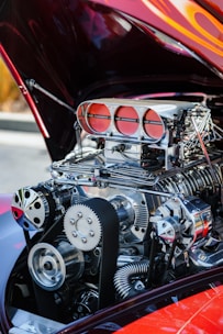A close-up view of a highly detailed, polished car engine with prominent metallic components and intricate parts. The engine is exposed with the car hood opened, and the surrounding area is slightly out of focus.