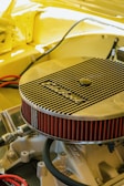 A detailed view of an engine component, specifically an Edelbrock air filter, installed on a car engine. The surrounding area is cast in a warm yellow hue, suggesting the car's body color. The angled shot highlights the filter's ribbed metallic cover and its bright red pleated edges. Various hoses and metal parts are visible under the hood, creating a sense of inner workings and mechanical intricacy.