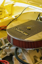 A detailed view of an engine component, specifically an Edelbrock air filter, installed on a car engine. The surrounding area is cast in a warm yellow hue, suggesting the car's body color. The angled shot highlights the filter's ribbed metallic cover and its bright red pleated edges. Various hoses and metal parts are visible under the hood, creating a sense of inner workings and mechanical intricacy.