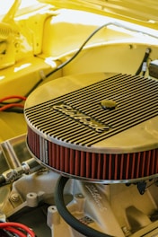 A detailed view of an engine component, specifically an Edelbrock air filter, installed on a car engine. The surrounding area is cast in a warm yellow hue, suggesting the car's body color. The angled shot highlights the filter's ribbed metallic cover and its bright red pleated edges. Various hoses and metal parts are visible under the hood, creating a sense of inner workings and mechanical intricacy.