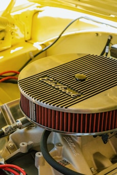 A detailed view of an engine component, specifically an Edelbrock air filter, installed on a car engine. The surrounding area is cast in a warm yellow hue, suggesting the car's body color. The angled shot highlights the filter's ribbed metallic cover and its bright red pleated edges. Various hoses and metal parts are visible under the hood, creating a sense of inner workings and mechanical intricacy.