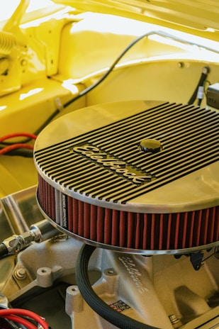 A detailed view of an engine component, specifically an Edelbrock air filter, installed on a car engine. The surrounding area is cast in a warm yellow hue, suggesting the car's body color. The angled shot highlights the filter's ribbed metallic cover and its bright red pleated edges. Various hoses and metal parts are visible under the hood, creating a sense of inner workings and mechanical intricacy.