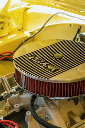 A detailed view of an engine component, specifically an Edelbrock air filter, installed on a car engine. The surrounding area is cast in a warm yellow hue, suggesting the car's body color. The angled shot highlights the filter's ribbed metallic cover and its bright red pleated edges. Various hoses and metal parts are visible under the hood, creating a sense of inner workings and mechanical intricacy.