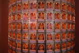 A cylindrical structure is densely covered with rows of small, ornate niches. Each niche contains a small, golden statue of a deity, illuminated with a warm orange light. The niches are decorated with intricate designs and inscriptions or labels in a script resembling Chinese characters.