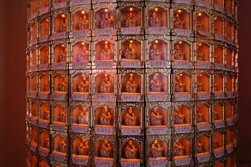 A cylindrical structure is densely covered with rows of small, ornate niches. Each niche contains a small, golden statue of a deity, illuminated with a warm orange light. The niches are decorated with intricate designs and inscriptions or labels in a script resembling Chinese characters.