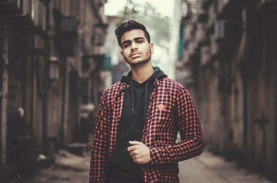 A young man stands confidently in an urban alleyway. He wears a black hoodie under a red and black checkered jacket. The background is blurred, highlighting old buildings or structures that create a narrow path.