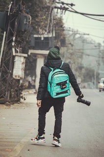 A person stands on a deserted street holding a camera with a long lens. The individual is wearing a black jacket, green hood, and possesses a turquoise backpack. They are looking towards the background, seemingly observing or capturing the urban surroundings, which include power lines and greenery.