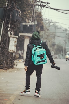 A person stands on a deserted street holding a camera with a long lens. The individual is wearing a black jacket, green hood, and possesses a turquoise backpack. They are looking towards the background, seemingly observing or capturing the urban surroundings, which include power lines and greenery.