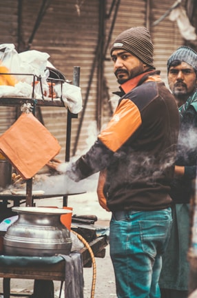 Two men are standing at a street food stall. One man is cooking with steam visible around him, while the other man looks towards the camera. Both are dressed warmly, wearing jackets and beanies. The stall has pots, cooking utensils, and is located on an urban street with a closed shutter in the background.