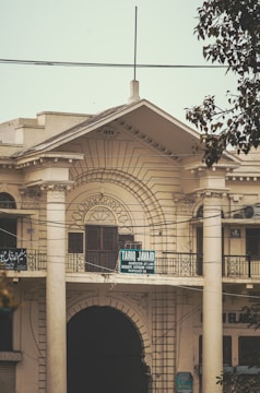 A historic building with decorative columns and ornate architectural details. The structure is marked with the year 1930 at the top. There is a signboard on the balcony, which reads 'Tariq Javaid, Barrister-at-Law, Advocate Supreme Court.' The facade features intricate designs and large archways, with some power lines visible in front. A tree partially obscures the view on the right side.
