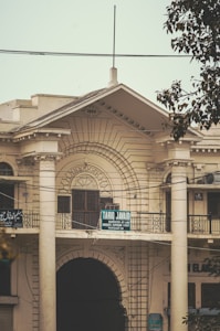 A historic building with decorative columns and ornate architectural details. The structure is marked with the year 1930 at the top. There is a signboard on the balcony, which reads 'Tariq Javaid, Barrister-at-Law, Advocate Supreme Court.' The facade features intricate designs and large archways, with some power lines visible in front. A tree partially obscures the view on the right side.