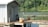 A wooden dock extends over a calm body of water, with a small bait shop or shack on the left. There are two large clay pots on the dock, and a heron stands on the edge of the water beside a bench. Dense green trees form a backdrop across the water.