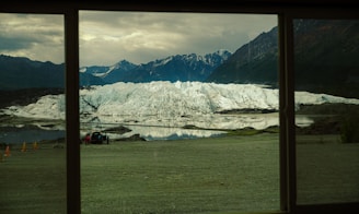 A large glacier with rugged, icy terrain surrounded by mountains and reflected in a still body of water is visible through a window. In the foreground, there are people near a parked vehicle and some orange cones on a grassy area.