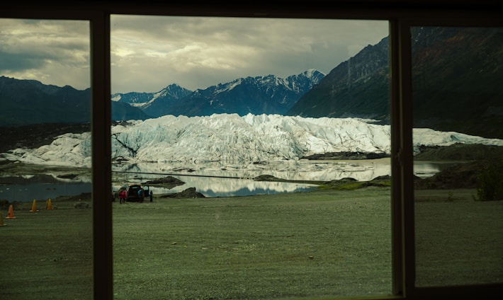 A large glacier with rugged, icy terrain surrounded by mountains and reflected in a still body of water is visible through a window. In the foreground, there are people near a parked vehicle and some orange cones on a grassy area.