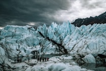 A group of people stands on the ice near an expansive and rugged glacier. The glacier features sharp, jagged peaks with a striking pattern of light blue ice and dark, dirt-filled crevices. A small body of water at the base reflects the icy terrain, while dark clouds loom overhead, adding to the dramatic and imposing atmosphere.
