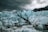 A group of people stands on the ice near an expansive and rugged glacier. The glacier features sharp, jagged peaks with a striking pattern of light blue ice and dark, dirt-filled crevices. A small body of water at the base reflects the icy terrain, while dark clouds loom overhead, adding to the dramatic and imposing atmosphere.