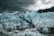 A group of people stands on the ice near an expansive and rugged glacier. The glacier features sharp, jagged peaks with a striking pattern of light blue ice and dark, dirt-filled crevices. A small body of water at the base reflects the icy terrain, while dark clouds loom overhead, adding to the dramatic and imposing atmosphere.