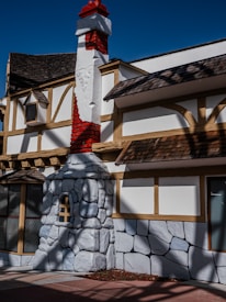 The image features a building with a rustic architectural style. Prominent wooden beams frame cream-colored walls, and a distinctive red brick chimney extends above the roofline. The base of the chimney is adorned with large, stylized stone blocks. The roof has small, shingled eaves, and there are windows with grid panels and wooden frames visible.