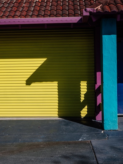A brightly colored garage door with a bold yellow hue is flanked by a teal pillar and a section of purple under the red tiled roof. A distinctive shadow pattern is cast on the door, creating a geometric design.