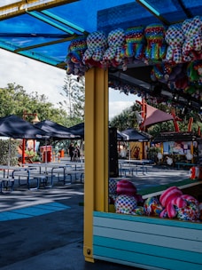 A colorful amusement park setting with stuffed toys hanging from a stall. Empty picnic tables are visible under black canopies, surrounded by trees and various small structures.