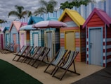 A row of colorful beach huts with striped deck chairs arranged in front. The huts are painted in a variety of pastel shades, including yellow, blue, pink, and orange. Palm trees can be seen in the background, and there is a striped beach umbrella among the deck chairs.