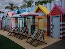 A row of colorful beach huts with striped deck chairs arranged in front. The huts are painted in a variety of pastel shades, including yellow, blue, pink, and orange. Palm trees can be seen in the background, and there is a striped beach umbrella among the deck chairs.