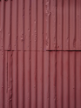 Corrugated metal sheets painted in a faded reddish-brown color with visible seams and texture. Each sheet is vertically aligned with visible rust spots and dents, giving a rugged, industrial appearance.