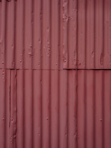Corrugated metal sheets painted in a faded reddish-brown color with visible seams and texture. Each sheet is vertically aligned with visible rust spots and dents, giving a rugged, industrial appearance.