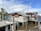 A series of wooden houses built on stilts over muddy water, with corrugated metal and palm leaves as roofing materials. A palm tree and a utility pole are visible near the structures. The sky is partly cloudy, creating a mix of shadows and light in the scene.