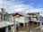 A series of wooden houses built on stilts over muddy water, with corrugated metal and palm leaves as roofing materials. A palm tree and a utility pole are visible near the structures. The sky is partly cloudy, creating a mix of shadows and light in the scene.