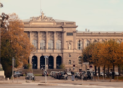 The image displays a grand historical building with classical architecture, featuring a detailed facade with large columns and arched windows. The name 'Université de Strasbourg' is visible on the building. The scene includes a number of parked cars in front of the building, a few people walking, and trees with autumn-colored leaves lining both sides.