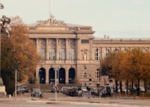 The image displays a grand historical building with classical architecture, featuring a detailed facade with large columns and arched windows. The name 'Université de Strasbourg' is visible on the building. The scene includes a number of parked cars in front of the building, a few people walking, and trees with autumn-colored leaves lining both sides.