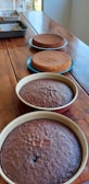 Four round cakes, two chocolate and two vanilla, rest on a wooden table. The cakes are in various stages of preparation. The chocolate cakes are in baking pans at the forefront, while the vanilla ones are placed on blue plates. In the background are additional items on a metal tray.