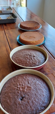Four round cakes, two chocolate and two vanilla, rest on a wooden table. The cakes are in various stages of preparation. The chocolate cakes are in baking pans at the forefront, while the vanilla ones are placed on blue plates. In the background are additional items on a metal tray.