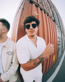 A young man with sunglasses and tattoos on his left arm is standing next to a rust-colored shipping container, wearing a white shirt and holding a cigarette. Another person is partially visible, facing away and wearing a jacket with the words 'The Stronghold' printed on it.